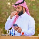 Man wearing keffiyeh drinking tea outdoors, using cellphone in Hafar Al Batin.